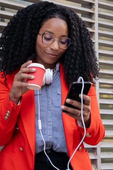 Black business woman with coffee in hand and headphones hanging around her neck, watching news with her mobile phone.