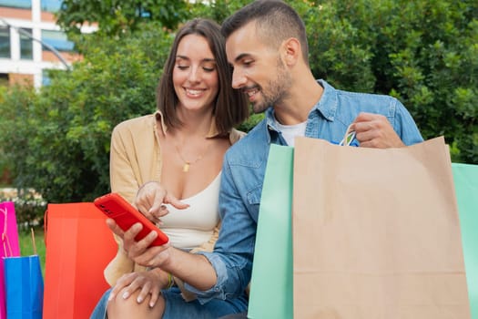 Pretty Caucasian couple of man and woman sitting holding shopping bags, browsing smartphone apps shopping online
