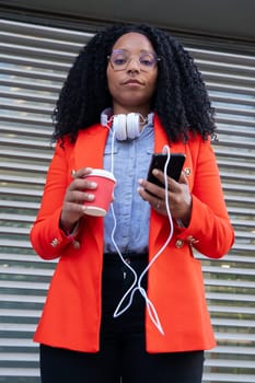 Serious black businesswoman with headphones, smartphone and coffee in hands outdoors.