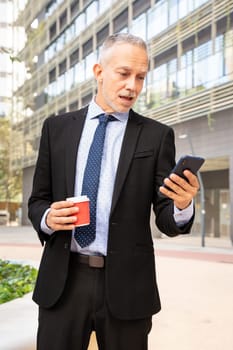 Satisfied with the results, the businessman walks down the street outside the office building, a mature boss holds a phone and coffee in his hands, writes messages and reads news online, using an app.