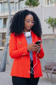 Black woman with headphones browsing the internet on social networks with mobile phone outdoors.