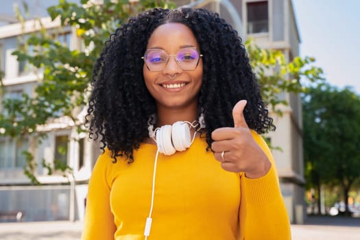 Close-up of happy curly haired young woman with glasses pointing thumb up. Facial expression of joy.