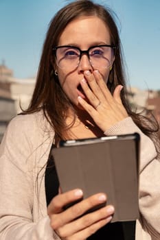 Beautiful young adult caucasian woman wearing glasses with surprise face with tablet pc looking at camera