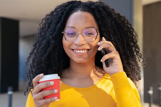 Young smiling black student talking on mobile phone while drinking coffee on university campus. Concept: university life