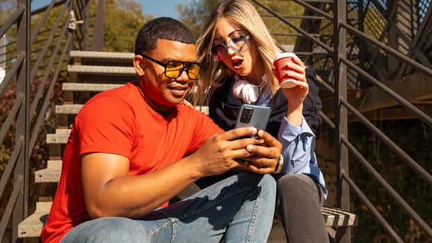 Happy diverse friends in the park sitting on a bench together looking at social media on smartphones talking and smiling.