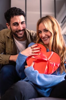 Beautiful young couple at home enjoying time together while celebrating Valentine's Day with a heart-shaped gift box in hand.