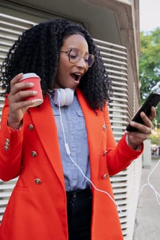 black business woman with coffee in her hand and headphones hanging around her neck, receiving good news on the mobile phone.vertical photography