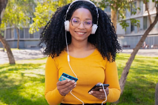 happy black woman wearing headphones pays with credit card on smartphone. Looking to the camera. Concept: online store payment