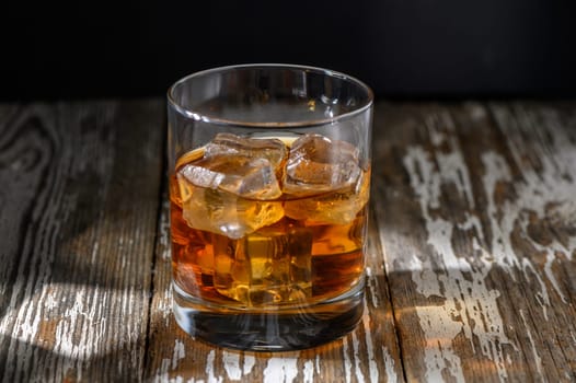 A single glass of whiskey sits on a weathered wooden surface, filled with ice and catching the warm glow of evening.
