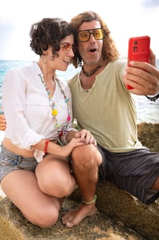 Front view woman and man wearing sunglasses on vacation by the sea laughing greeting by video call to a family member.Vertical