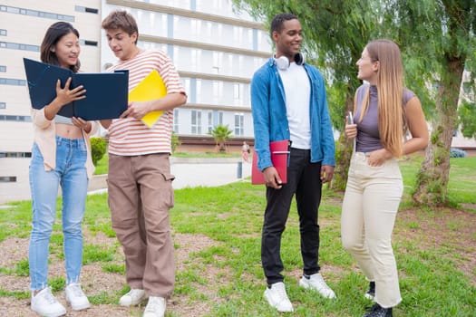 Happy university student friends talking and laughing while reviewing syllabus notes on campus. Diverse Asian and European youth