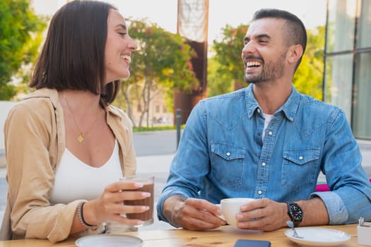 Two young friends chatting on a cafeteria terrace drinking coffee, laughing and having a good time after work.