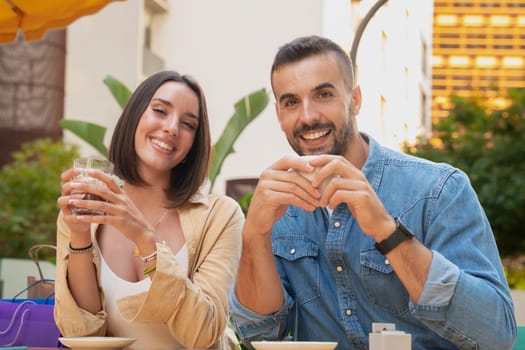 Young couple sitting with shopping bags on a terrace, looking at the camera with a coffee and smiling