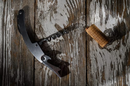 A sleek corkscrew lies beside a wine cork on aged wooden planks, illuminated by soft natural light from above.