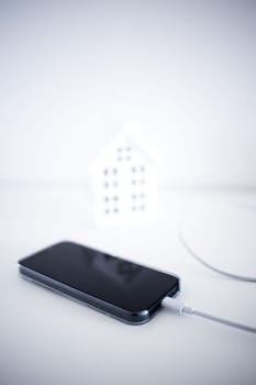 Smartphone charging by miniature house on white background.