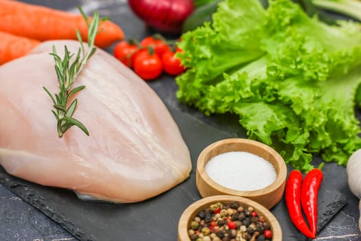 Chicken diet breast with a sprig of rosemary on top lying surrounded by herbs, seasonings and vegetables on a dark background close-up. The concept of healthy eating, dietary and natural products, cooking. High quality photo