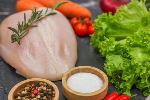 Close-up of raw chicken fillet with a sprig of rosemary lying surrounded by vegetables, herbs, spices on a dark background. The concept of diet, detox, proper and healthy nutrition, recipes. High quality photo