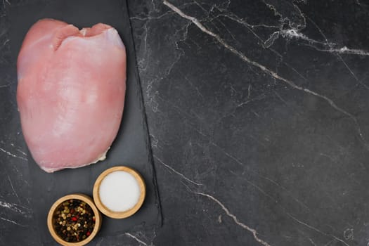 Close-up of raw turkey fillet lying on a black stone plank with spices and salt in wooden bowls side by side on a dark background with space for text. The concept of proper and healthy nutrition, diet, menu, natural and organic products. High quality photo