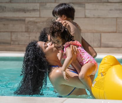 Mother kissing daughter in swimming pool with family playing in background
