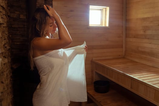 A young woman relaxes and sweats in a hot sauna wrapped in a towel. girl in the sauna. Finnish sauna interior, classic wooden sauna with hot steam. Russian bath