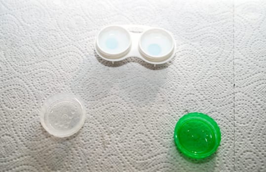 Container for storing lenses and contact lenses. The container is white with green, there are lenses in it. The table is covered with a paper towel for sterility at home. The container is open, you can see the lenses in it.