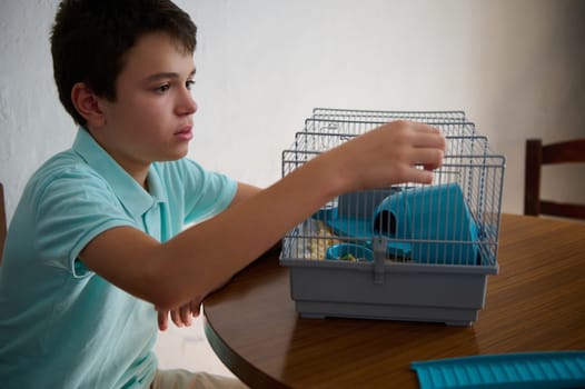 A young boy carefully looks after a pet in its cage while seated at a table. The scene captures his attentiveness and love for his animal friend.