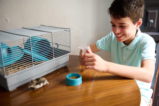 A cheerful boy sits at a table with his hamster and its cage, enjoying quality time together. The scene captures joy and care between a pet owner and his adorable, furry friend.