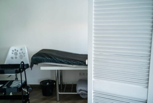 table for massage and other cosmetology services. covered with a blanket and a disposable sheet. next to it is a white chair. in the foreground is a white screen.