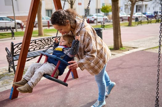 Loving parent spending quality time with their child on a swing at a public park, surrounded by nature and urban scenery. A tender moment of care, joy, and family bonding.