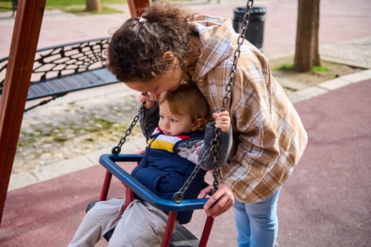 A mother enjoys a joyful moment with her young child, bonding as they play on a swing in a serene park setting, showcasing familial love, care, and outdoor activities.
