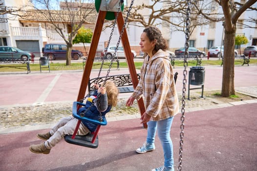 A mother gently pushes her child on a swing at a playground on a sunny day. The outdoor park setting features play equipment, surrounding trees, and a cheerful scene of togetherness and connection.