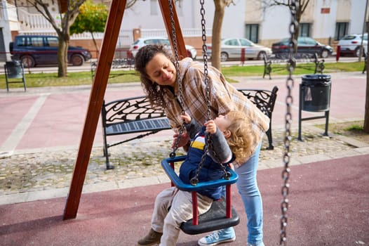 A loving mother interacts with her young child on a swing in a sunny playground. The joyful moment reflects family bonding, play, and outdoor activities, emphasizing love, care, and togetherness.