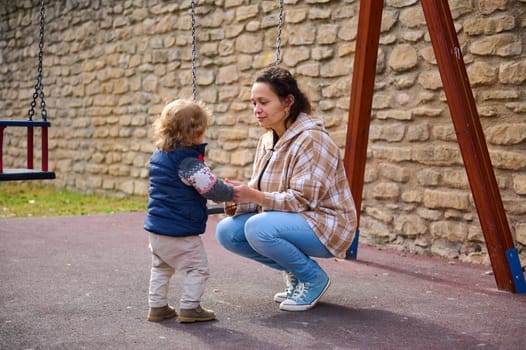 A mother kneels as she interacts with her young child at a sunny playground. The surrounding creates a joyful and familial scene, highlighting tenderness and parenting in an outdoor space.