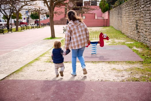 A mother and her young child walking hand-in-hand in a park playground during a sunny day. The scene captures a parent-child bond and outdoor activity.