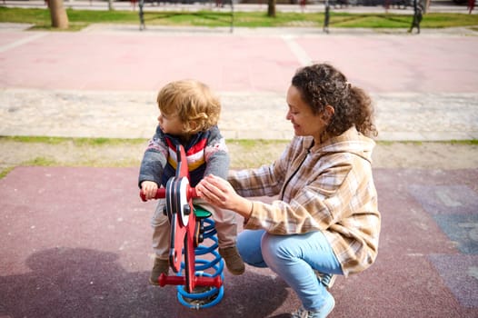 A joyful moment of a woman and toddler playing at a colorful playground. The child is riding a spring-mounted toy as the woman supports them with visible enjoyment.