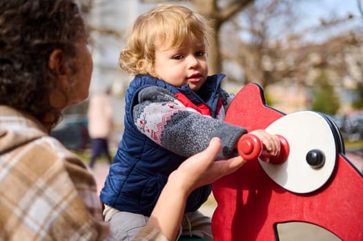 A loving mother assists her young child on a playground toy, capturing a joyful and engaging moment outdoors. The image evokes themes of family, bonding, and playful innocence in a natural setting.