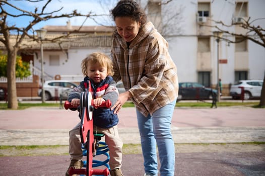 A cheerful moment of a mother helping her young child ride a playground toy in a sunny park. This image captures family bonding and outdoor fun.