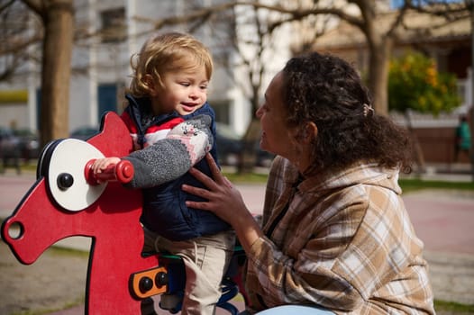 A joyful scene showcasing a loving mother and her young child enjoying a sunny day at the playground. The child is riding a red rocking horse while the mother supports and guides them.