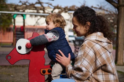 A mother lovingly supervises her child riding a playground rocking horse. The location is a park, evoking fun, care, and family connection under afternoon sunlight.