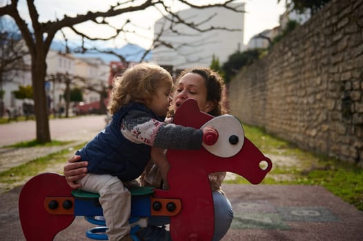 A mother and her young child spend quality time playing on a toy horse in a scenic park. The serene atmosphere and joyful interaction reflect familial love and life moments.
