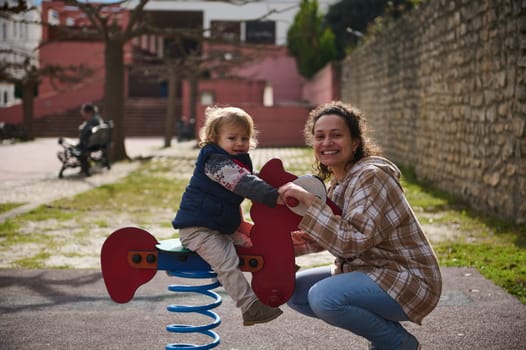 A cheerful mother and her young child enjoy playing on a spring rider at a park. The sunny outdoors setting highlights the joy and interaction during their playtime.