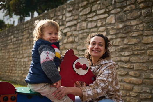 A cheerful mother and her smiling young child enjoy playful moments together in the park on a sunny day. Their happiness radiates as they embrace a joyful outdoor activity surrounded by nature.