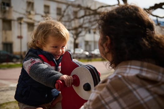 A young child rides a red playground toy, smiling while a mother watches nearby, enjoying outdoor bonding time in a warm and sunny urban park setting.