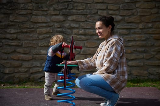 A mother is interacting with her child while they play on a spring rider toy at an outdoor playground. The moment reflects joy, bonding, and family fun in a relaxed setting.