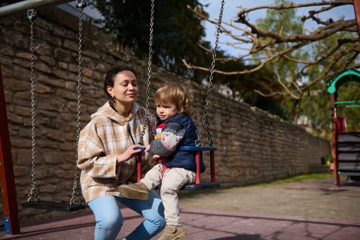 A mother and her young son bonding together in a park while using a swing on a sunny day.
