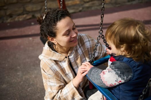 A mother lovingly interacts with her young child who is seated on a swing outdoors, portraying warmth and togetherness in a sunny, leisurely setting.