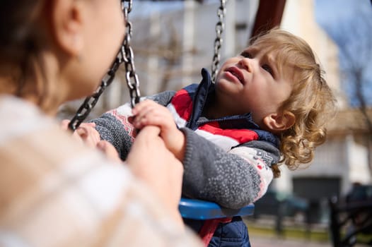 A child on a swing set holds hands with an adult in a sunny outdoor playground.