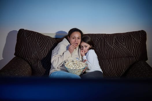 A mother and daughter sit on a cozy sofa, sharing popcorn while watching a movie, emotions ranging from excitement to suspense, creating a relaxed family bonding time.