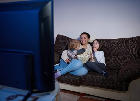 A mother and her children enjoying quality time together while watching TV and eating popcorn. This cozy scene highlights family bonding and relaxation at home during a leisurely evening.