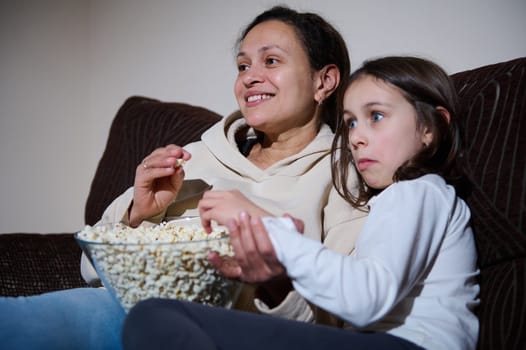 A mother and her daughter sitting on a couch, eating popcorn, and enjoying a movie night together, showcasing family bonding and quality time spent watching a film.